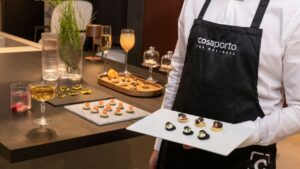 Un cameriere con grembiule "Cosaporto for Business" presenta un vassoio di finger food gourmet, parte del servizio "Cosaporto Digital Catering", circondato da eleganti drink e antipasti su un tavolo raffinato.